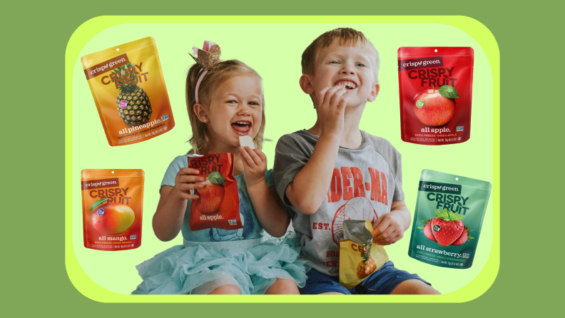 Two children smile with mouths open enjoying freeze dried fruits together while surrounded by four bags of freeze dried fruits.