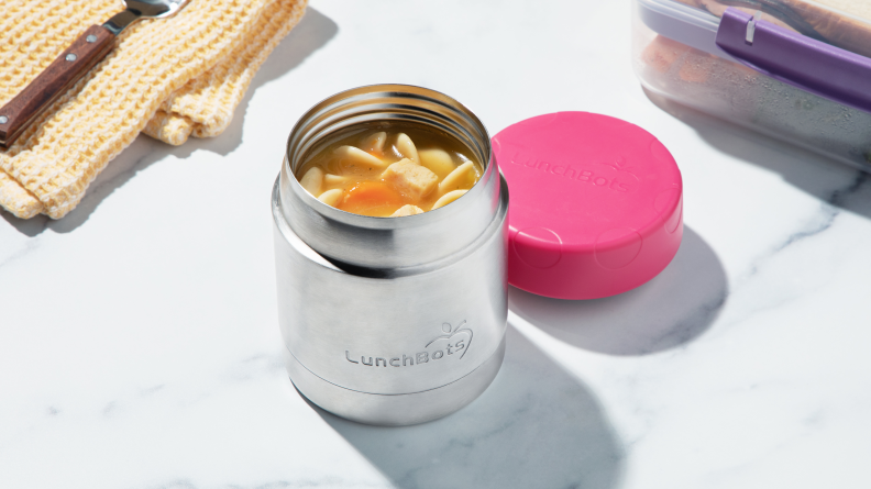 A stainless steel thermos filled with chicken noodle soup sits on a kitchen counter. The pink lid for the thermos sits next to it.