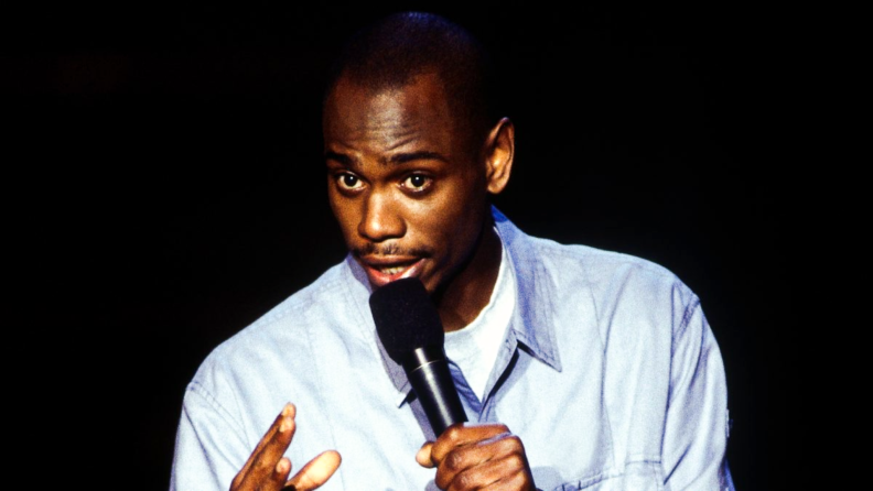 Comedian Dave Chappelle performs stand-up on stage, microphone in hand.