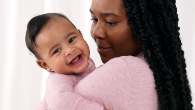 A smiling baby in a pink outfit rests happily on a woman’s shoulder, both dressed in matching pink.