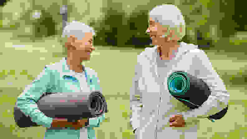 Two elderly women carrying yoga mats smile at each other while in conversation