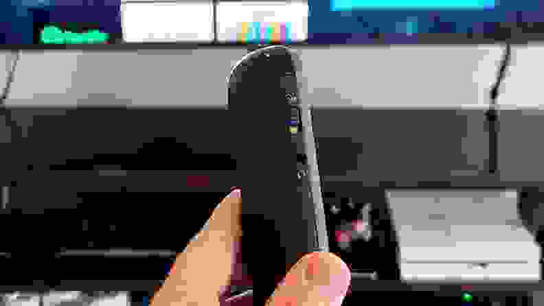 A hand holds the black, rounded remote in front of a TV console with a hands-free switch showing.
