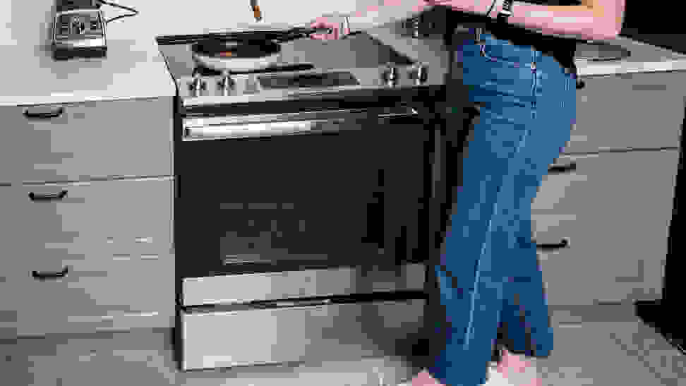 A person wearing jeans cooking food in a pan on an electric stovetop.