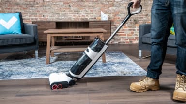 A man dressed in jeans and brown work boots uses a white vacuum on a wood floor next to a blue rug.