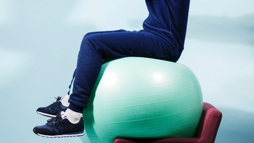 A performance artist balances on an exercise ball and a chair.