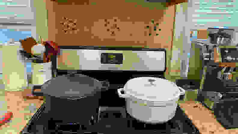 On a gas stovetop, on the left there's a black Perfect Pot with its lid on; to its right, there's a white Staub Dutch oven with its lid on. The Perfect Pot is taller than the white Dutch oven.