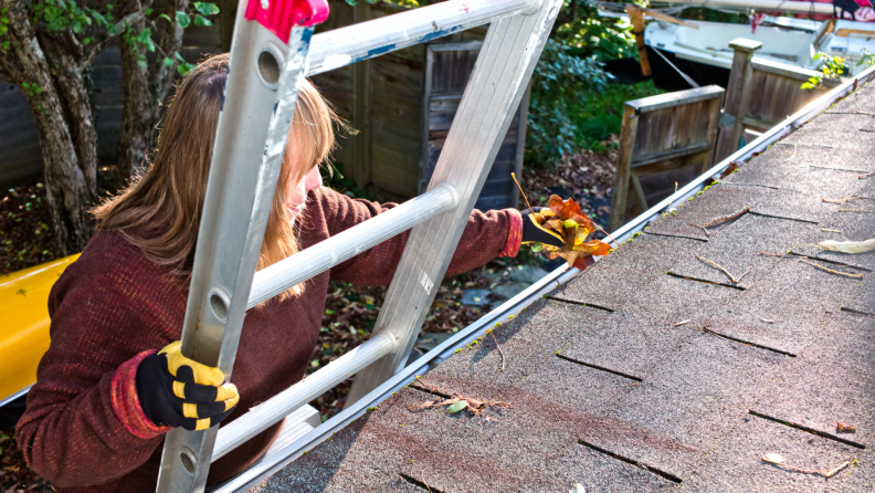 A woman standing on a ladder cleaning out a gutter that is full of leaves