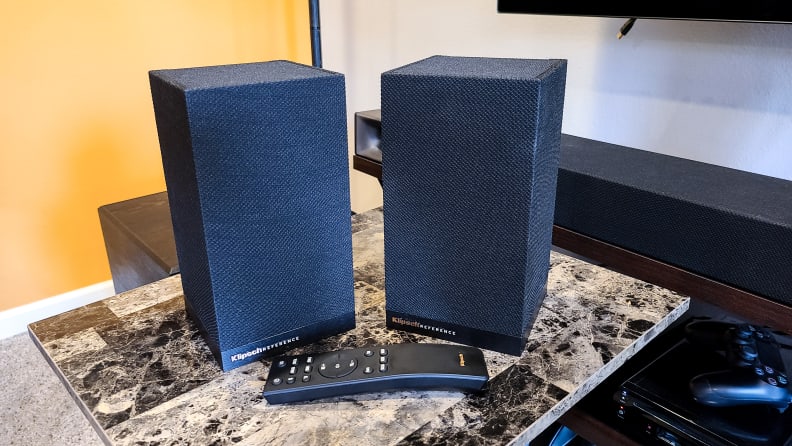 A pair of all-black speakers wrapped in acoustic fabric sit in front of a black, rubberized remote with white-lettered buttons that include subwoofer and surround sound control.