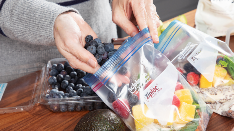 A person adds fresh blueberries into a freezer bag of smoothie ingredients.