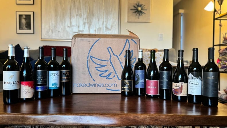 A row of wine bottles from Naked wine on top of wooden table surrounding a cardboard box.