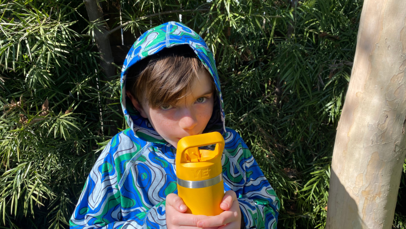 A child drinks from a yellow Yeti Rambler outside under trees.