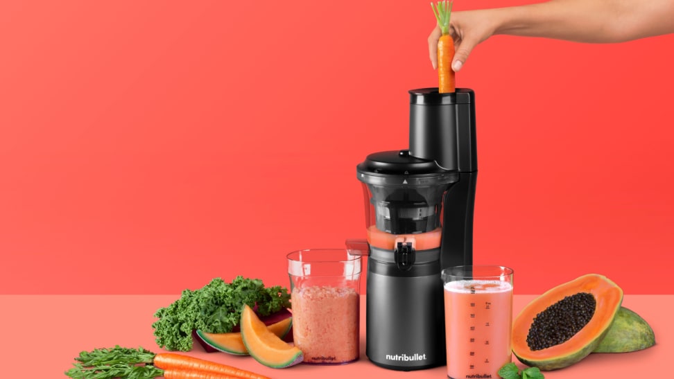 A NutriBullet Slow Juicer is in the center of the image. A person is pushing a carrot through the juicer. There are papayas, carrots, and melon scattered around the juicer.