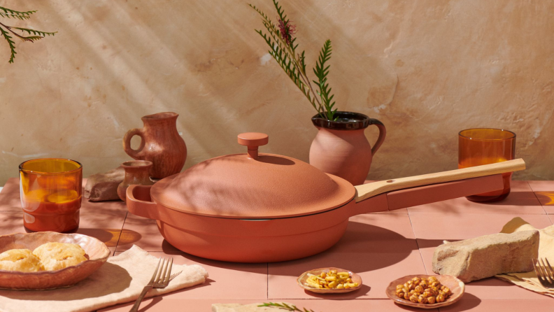 The terracotta-colored Always Pan is in the center of the image. The color is earthy with an orange hue. Next to the pan, pottery vases, drinkware, and plates of nuts and snacks are on display..