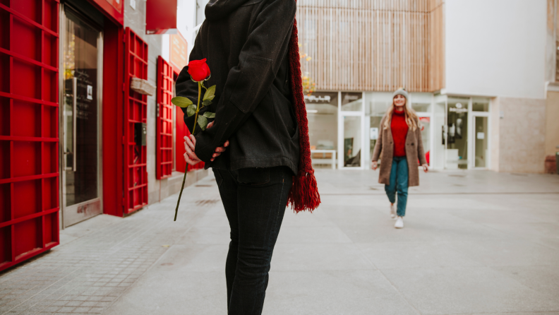 A person in a black coat and red scarf stands in an urban setting, holding a single red rose behind their back as they prepare to surprise their approaching partner, who is smiling and dressed in a cozy winter outfit.