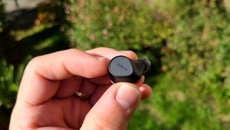 The black and silver Elite 7 Pro earbud is held in a hand in front of greenery.