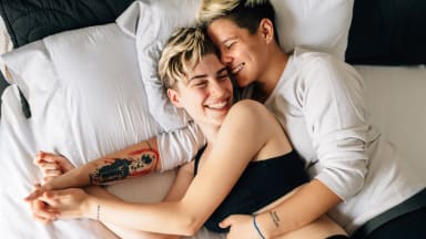 A queer couple cuddles in bed together, one spooning the other.