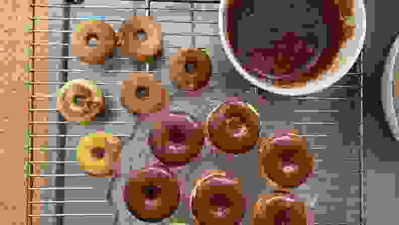 Unglazed mini donuts on a drying rack next to a pink glass tray filled with chocolate glazed mini donuts.