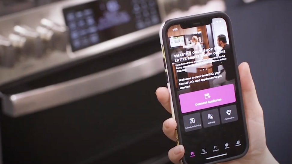 A person is holding a phone in front of a GE range. On the phone screen, it's the GE SmartHQ app that allows users to connect their WiFi-enabled appliances to their handsets.