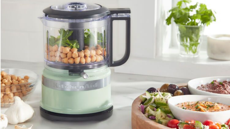 A small surf-green food processor sits on a countertop beside various dips, garlic bulbs, and so on.