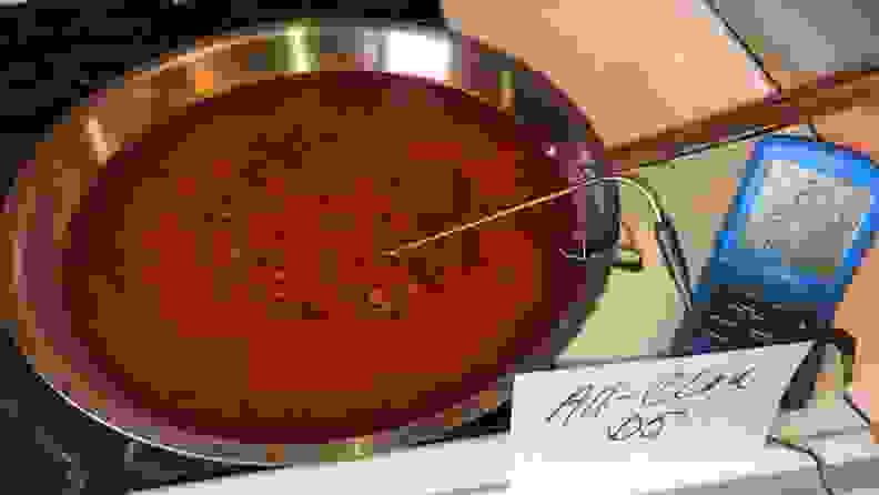 A probe thermometer is used to measure simmering tomato sauce inside a stainless steel pan.