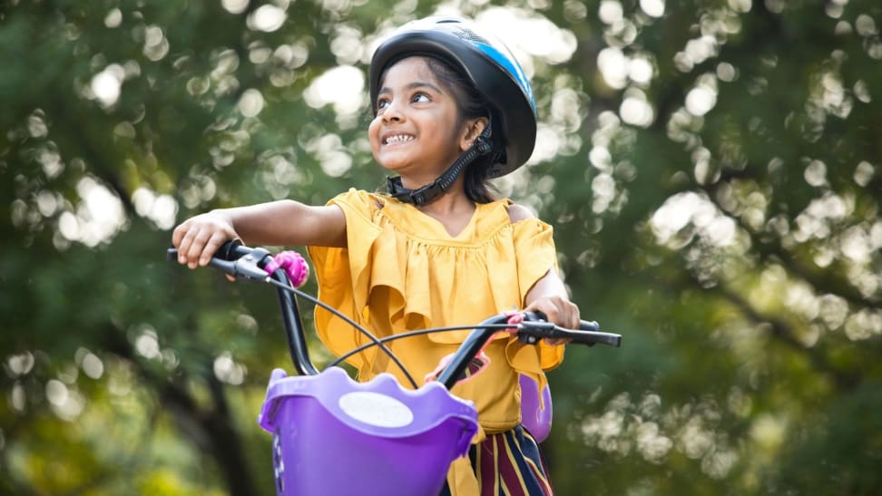 A girl beams while taking her bike for a ride outside. She's wearing a sunny yellow shirt and has a cute purple basket.