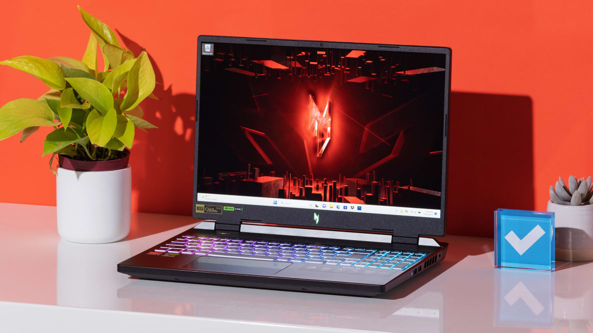 The Acer Nitro 16 sitting atop a table between a plant an the Reviewed logo.