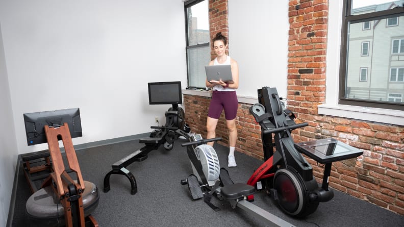 woman standing in a room with many different rowing machines and a laptop.