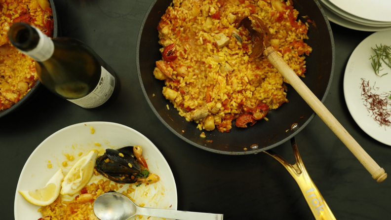 A pot full of cooked food in a black Frök pan is in the center of a black tabletop. Around the pan, some plates and a wine bottle are scattered around.