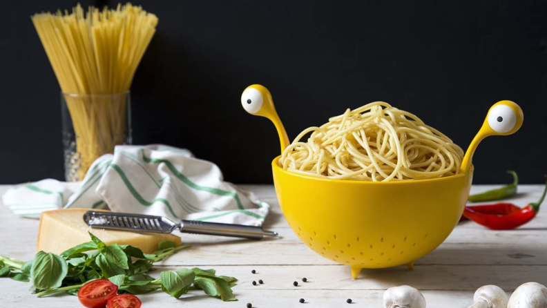 Yellow monster-inspired colander with two eye shaped handles on both sides with boiled spaghetti inside. Colander is next to uncooked white mushrooms, whole black peppercorns, red sliced tomatoes, a block of parmesan cheese, a grater, red peppers, and a white and green dish towel and uncooked pasta.