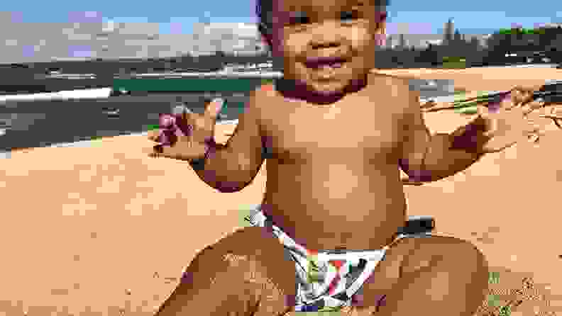 A baby wearing a Nageuret diaper sitting on a beach, smiling.