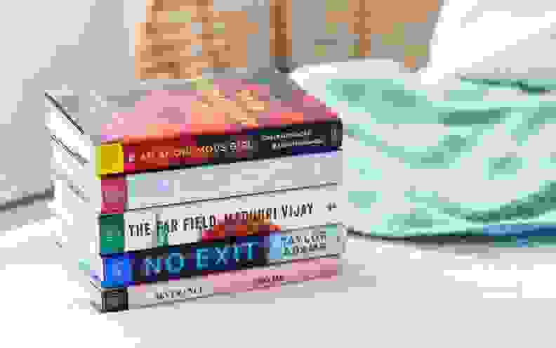 Five novels (from a Book of the Month subscription) are stacked on a bed.