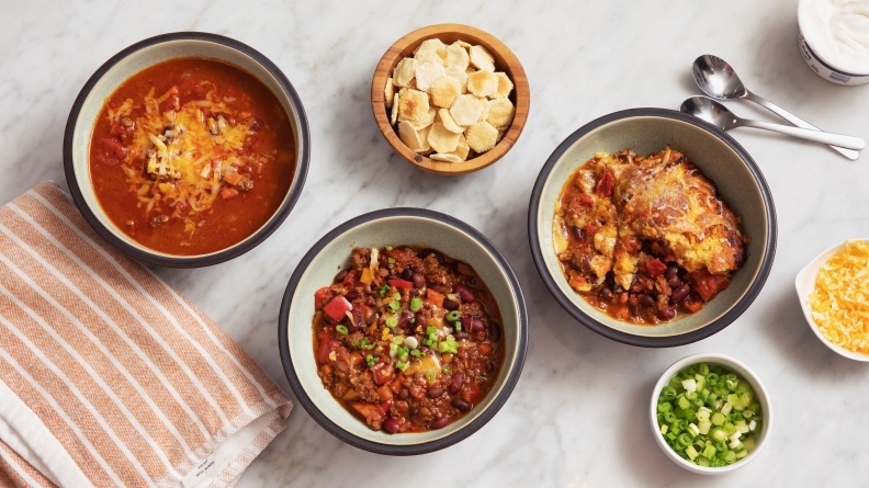 Three bowls of chili on a marble surface surrounded by toppings