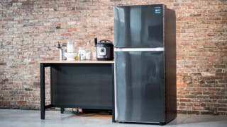 A top-freezer fridge stands in a room against a brick wall