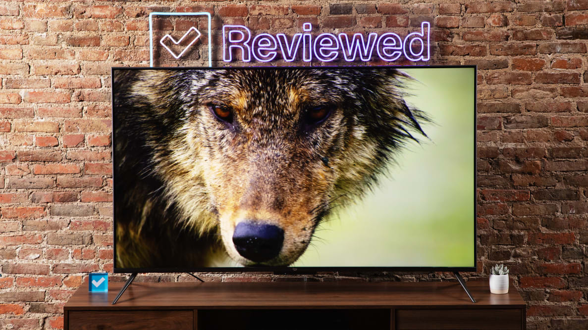 The 2022 Amazon Fire TV Omni sitting on a wooden table in front of a brick wall with a Reviewed neon sign displaying the image of a wolf.