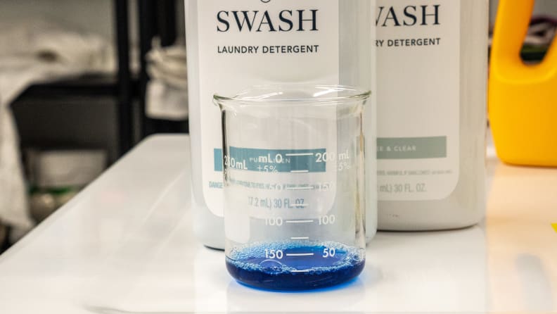 A beaker sits in front of two swash bottles, filled to a 25-milliliter line with blue detergent.