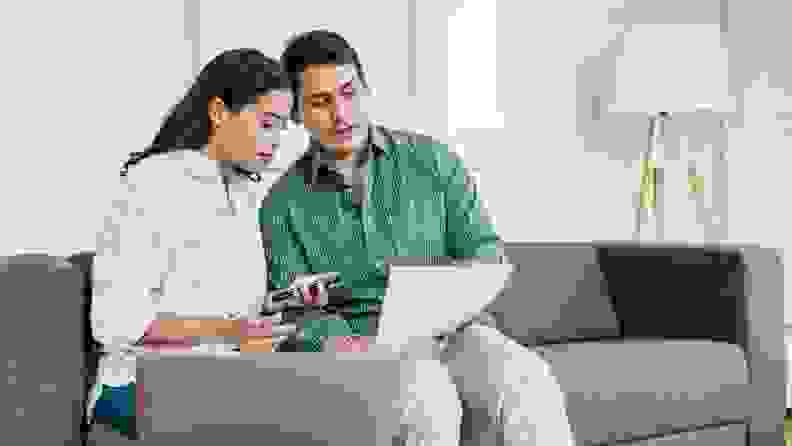 A couple sits on a couch as they look at a computer screen and documents
