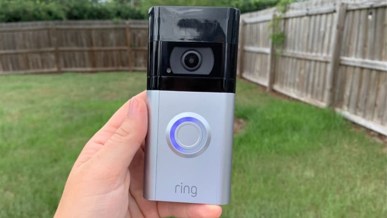 Person holding up a Ring Security Camera Doorbell is in front of yard outdoors.