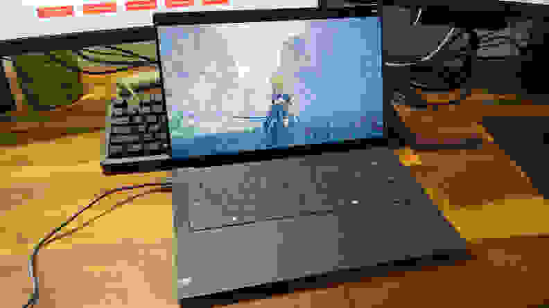 The Dell XPS 14 laptop opened to display a game on screen on a wooden desk in front of a desktop computer monitor.