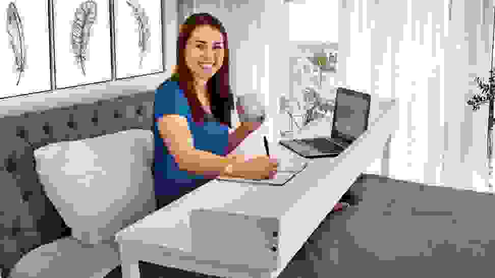 A smiling remote worker writes using an over-bed desk with a small laptop on it.