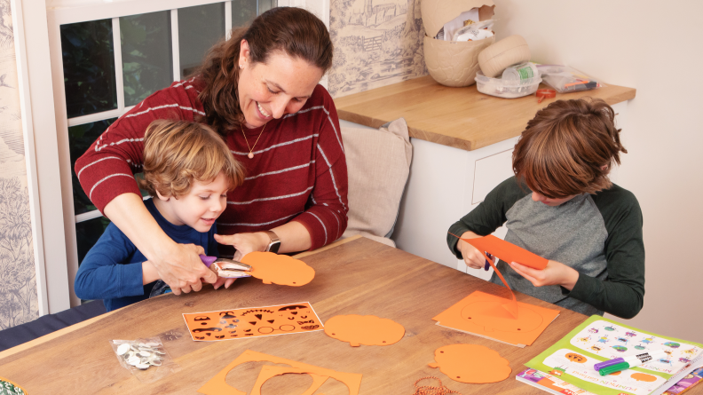 A parent helps a child do a pumpkin craft activity at a table while another child works independently.