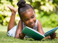 Young girl reading book outdoors in the grass while smiling