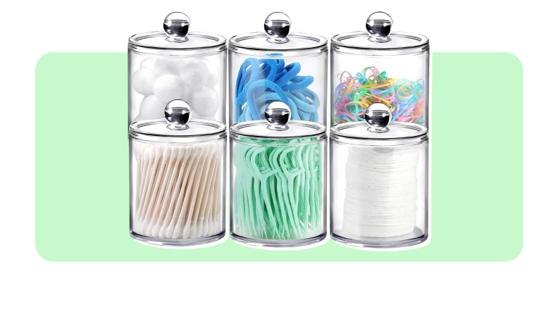 Six clear jars with lids filled with toothpicks, cotton pads, cotton balls, q-tips and hair scrunchies in front of light green background..
