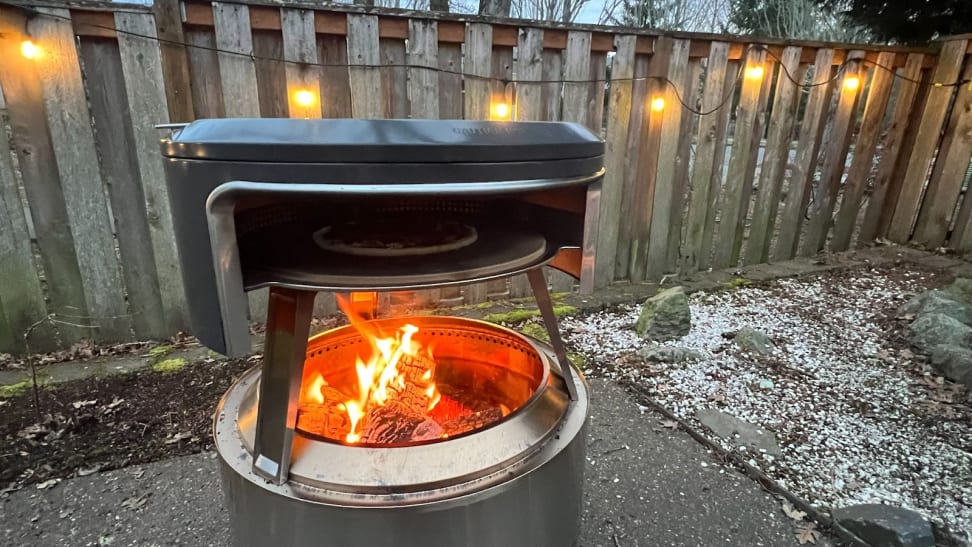 Solo Stove Pi Fire pizza oven with open flame outdoors in front of wooden fence with string lights.