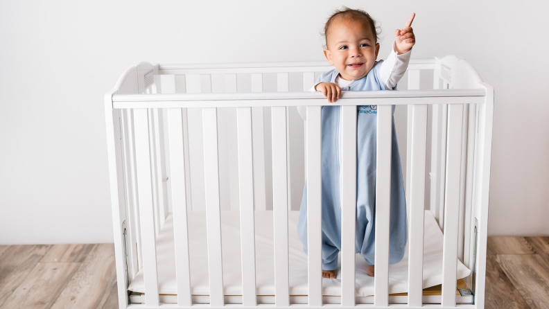 A baby stands inside a crib wearing a blue onesie.