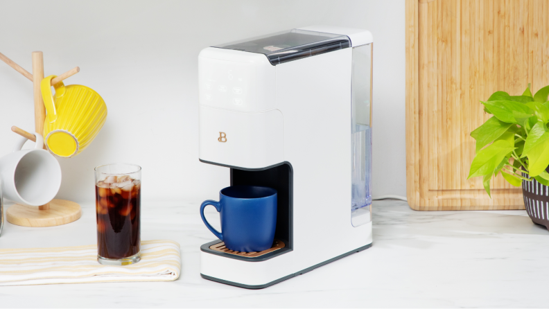 The coffee maker with a blue cup in it while a cup of iced coffee sits next to it.