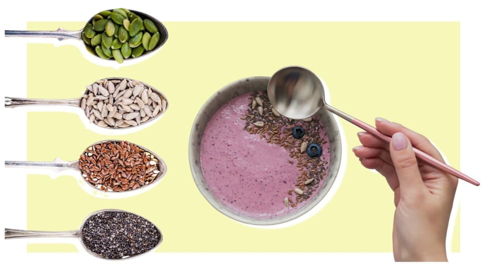 Four spoons hold different types of seeds next to person holding spoon above an açaí bowl.