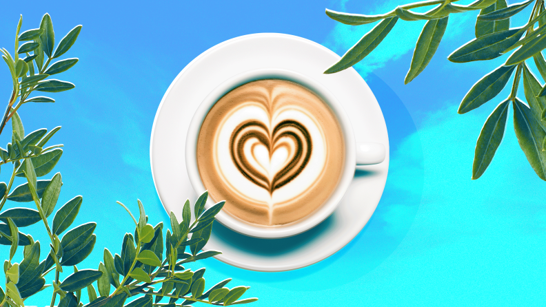 A latte showing heart latte art on a blue background surrounded by plants