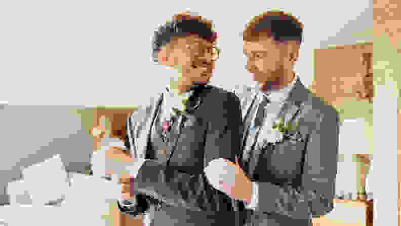 Married LGBTQ+ couple standing and looking at each other on their wedding day. They are dressed in suits and smiling.