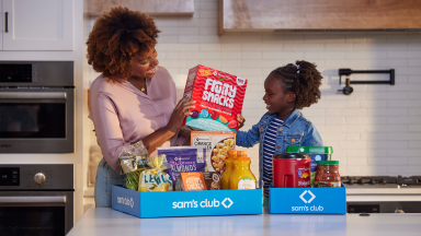 A mother and daughter smile while unpacking Sam's Club grocery boxes filled with snacks, drinks, and household essentials in a modern kitchen.