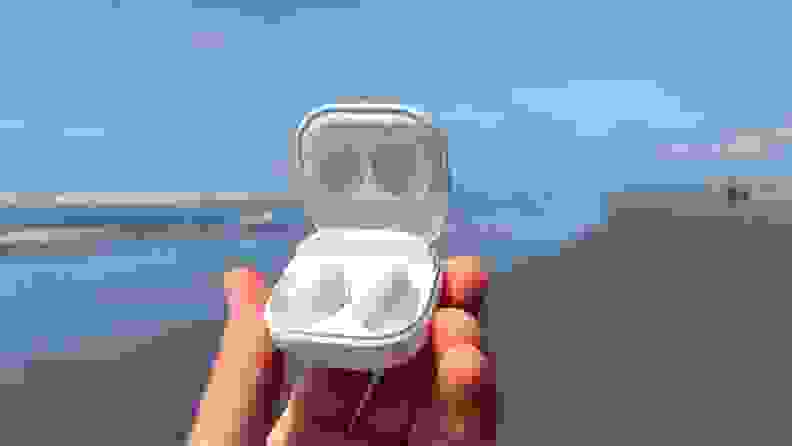 Samsung Galaxy Buds 2 are held in their white case with the micro USB showing against a sunny beach and a blue ocean.